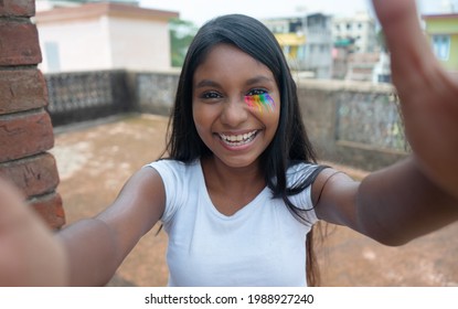 A playful dark skinned young girl of LGBTQ community with rainbow eye makeup laughing and taking a selfie | teen girl smiling cheerfully and giving hugs 
