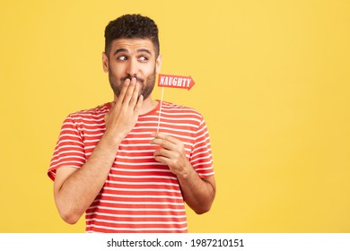 Playful disobedient man with beard in striped t-shirt shyly closing mouth with fingers holding sign on stick with naughty inscription. Indoor studio shot isolated on yellow background
