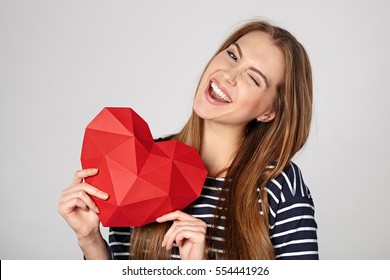 Playful emotional woman showing red polygonal paper heart shape winking at camera