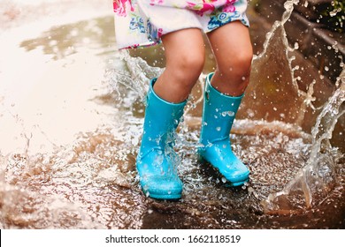 Playful excited preschool child in casual clothes and rubber boots with blue umbrella laughing and jumping in puddle smiling at camera while playing in park - Powered by Shutterstock