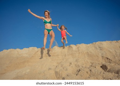 Playful family moment a woman and girl jumping from a sand ridge at the beach, with a backdrop of blue sky - Powered by Shutterstock