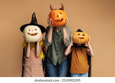 Playful family wearing costumes posing and having fun, holding carved pumpkin isolated on beige background, halloween celebration concept - Powered by Shutterstock