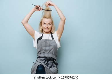 Playful fooling blonde female hairdresser in apron is pulling her bang up, making funny face over blue background. while sitting on a stool.