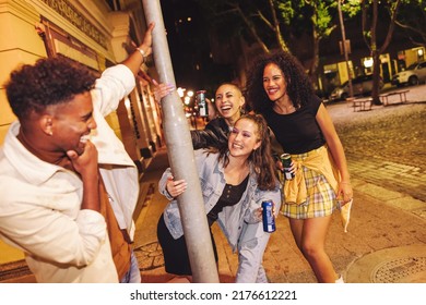 Playful friends having fun in the city at night. Group of carefree young friends laughing happily while standing around a pole. Multicultural young people some beers on the weekend.