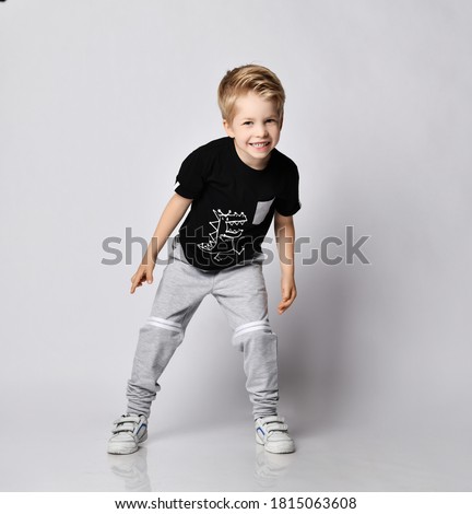 Playful frolic blond kid boy in sunglasses, black t-shirt with dinosaur print and gray pants stands leaning forwards going to run out playing catch-up over gray background