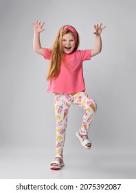 Playful, frolic redhead kid girl in pink t-shirt, colorful pants and sandals plays dino, monster roaring, holding hands with claws up and moving, walking straight towards camera
