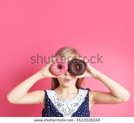 Playful girl holding donuts on her eyes. Close up portrait of a funny girl with long hair having fun with colorful donuts against her eyes. Satisfied child with a bandage on his hair, showing tongue.