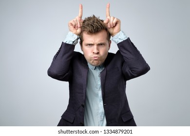 Playful Handsome Bearded Man In Suit Looking At Camera With Funny Face And Cow Horns Fingers. Indoor Studio Shot.