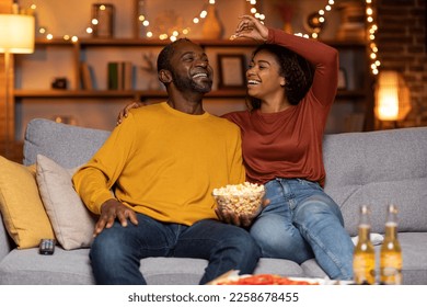 Playful happy beautiful adult african american lovers man and woman in casual having fun at cozy house in the evening, cheerful wife feeding her laughing husband with popcorn, copy space - Powered by Shutterstock