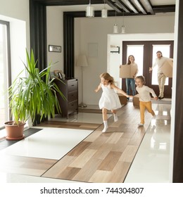 Playful happy kids running into new big own beautiful house, family moving in day concept, excited children exploring home interior having fun together, parents holding cardboard boxes at background 