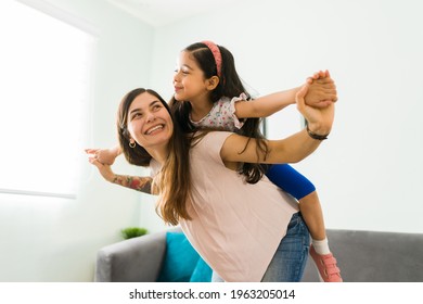 Playful happy mom giving a piggyback to her little happy daughter and playing to fly in the living room  - Shutterstock ID 1963205014