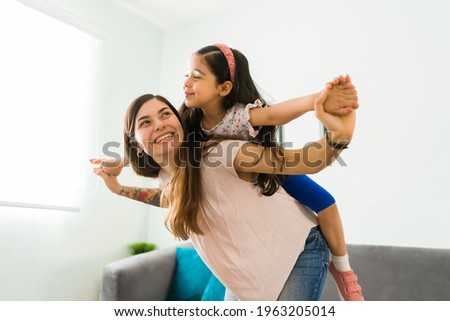Playful happy mom giving a piggyback to her little happy daughter and playing to fly in the living room 