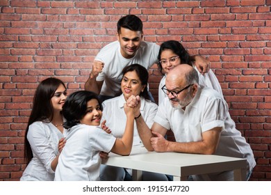 Playful Indian Asian Kid And Grandfather Arm Wrestling At Home With Other Family Members Looking, Wearing White Cloths