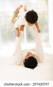 Playful Indian Father Lying On The Bed And Lifting Up His Little Son At Home