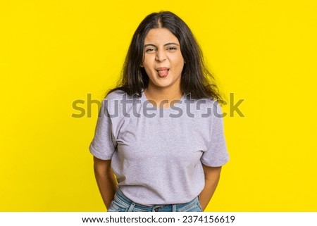 Playful Indian young woman showing tongue making faces at camera, fooling around, joking, aping with silly face, teasing, bullying, abuse. Cheerful funny Arabian girl isolated on yellow background