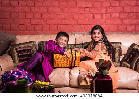 Playful Indian/Asian kids in ethnic wear with Diwali gifts, smiling or fighting for gifts while sitting on Sofa