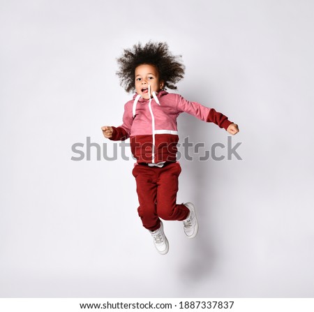 Playful little African American woman in stylish modern red jumpsuit jumps high on a gray background. Curly preschool girl having fun on a gray background. The concept of comfortable baby clothes.