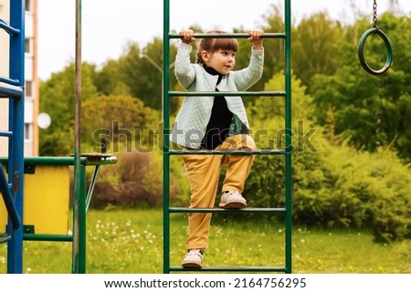 A playful little baby girl climbed an iron ladder on an outdoor playground on a sunny day. A cheerful little girl climbs the stairs on the playground. Games for the development of children outdoors.