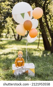 Playful Little Boy In Stylish Wear Standing Inside Wicker Basket Decorated With Colorful Helium Balloons. Birthday Celebration Of Little Child On Fresh Air.