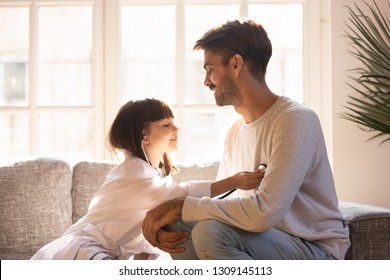 Playful Little Daughter Playing Funny Game As Doctor Having Fun With Dad Sitting On Couch, Cute Child Girl Pretending Nurse In Medical Uniform Holding Stethoscope Listening To Father Patient At Home