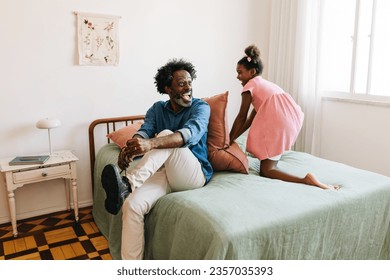 Playful little girl tossing a pillow towards her father while he ties his shoelaces. Happy black girl having fun with her dad in the morning, starting his work day with a cheerful mood. - Powered by Shutterstock