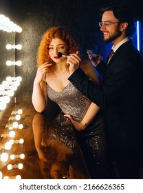 Playful Male Makeup Artist Tickles Nose Female. Curly Red Haired Woman In Evening Dress And Handsome Bearded Man In Suit Near Big Mirror In Dressing Or Make Up Room. Couple Before Going To Party