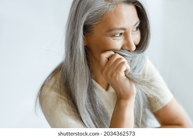 Playful Middle Aged Asian Woman Hides Face With Beautiful Grey Hair Sitting On Light Background In Studio. Mature Beauty Lifestyle