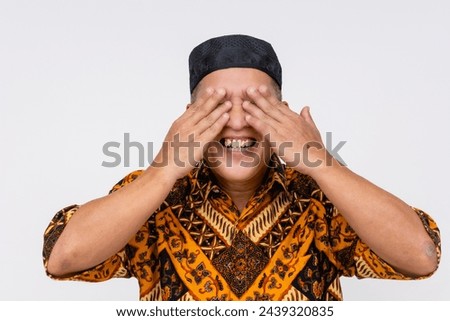 Playful middle aged Indonesian man in traditional batik attire and kopiah hat covering his eyes. Isolated on white.