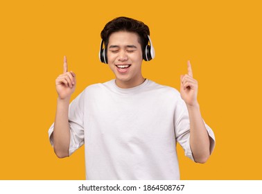 Playful Mood. Cheerful Asian Man Listening To Music In Wireless Headphones With Closed Eyes, Dancing And Raising Fingers Up, Posing Isolated On Orange Studio Background, Enjoying Favorite Song - Powered by Shutterstock