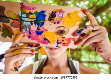 Spielerisches Porträt einer jungen, wunderschönen Künstlerin, die mit Farbe bedeckt ist und durch die Farbpalette ihres Malers die Kamera anschaut und lächelt. Kreativität und Individualität.