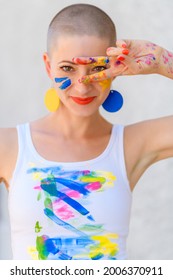 Playful portrait of a young gorgeous female artist painter covered in paint, looking and smiling at camera through her fingers. Creativity and individuality concept.