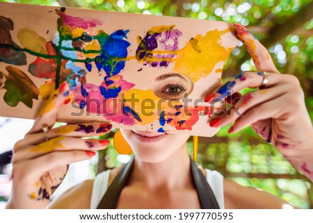 Playful portrait of a young gorgeous female artist painter covered in paint, looking and smiling at camera through her painter's palette. Creativity and individuality concept.