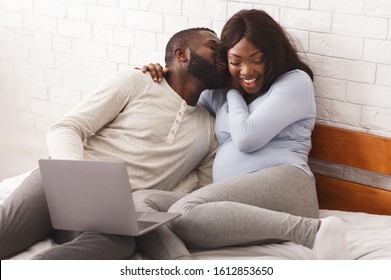 Playful pregnant african woman and her husband laying on bed, using laptop, man whispering words of love - Powered by Shutterstock