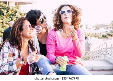 Playful Real Life Concept With Three Cheerful Smiling Middle Age Women Friends Anejoying And Laughing Togehter Playing With Soap Bubbles In Outdoor Leisure Activity - Playful And Happiness Concept