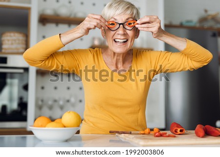 Playful senior woman holding slices of  vegetables over her eyes  and having fun in the kitchen