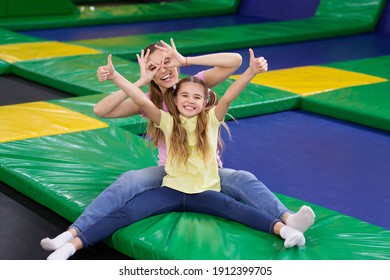 Playful Silly Mom With Her Teen Daughter Sitting At Trampoline Area Of Entertainment Centre, Showing Thumbs Up Gesture. Mother And Her Child Recommending To Visit Indoor Kids Playground