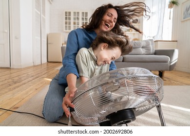 Playful Single Mother Bonding With Son Excited Playing Together At Home With Big Fan Blowing Cool Wind In Living Room. Mum And Child Enjoy Time Together At Conditioning Ventilator During Summer Heat