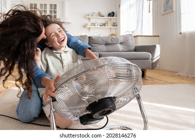 Playful Single Mother Bonding With Son Excited Playing Together At Home With Big Fan Blowing Cool Wind In Living Room. Mum And Child Enjoy Time Together At Conditioning Ventilator During Summer Heat