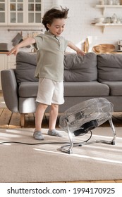 Playful Small Kid Alone In Living Room Enjoy Fresh Cool Breeze Blowing From Big Industrial Retro Ventilator Or Air Conditioner. Cheerful Kid Has Fun Indoors With Strong Cold Wind In Hot Summer Season