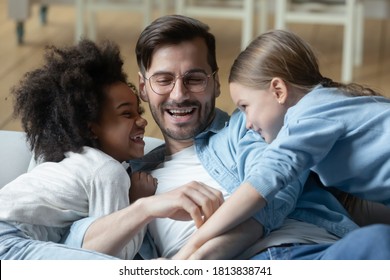 Playful Small Little Mixed Race Children Daughters Tickling Laughing Young Father, Having Fun Together On Sofa. Emotional European Daddy Enjoying Spending Weekend Playtime With Energetic Diverse Kids.