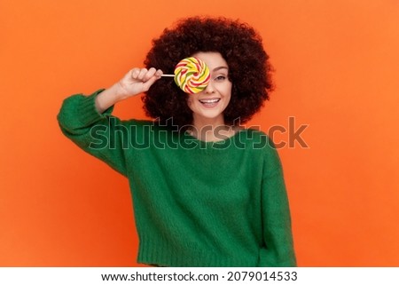 Playful woman with Afro hairstyle wearing green casual style sweater covering mouth with multicolor lollipop, having fun, childish behavior. Indoor studio shot isolated on orange background.