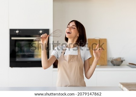 Playful woman in apron singing a song, pretending whisk to be a microphone, having fun and fooling around in modern kitchen interior, empty space.
