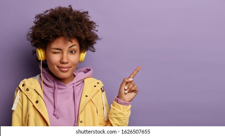 Playful woman blinks eyes, has appealing look, curly hairstyle, points index finger on copy space attracts your attention to advert listens music in playlist with modern headphones wears casual outfit
