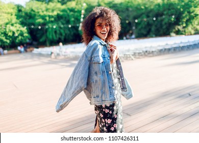 Playful woman wraps herself over her shoulder and looking in camera. Wearing. Stylish hipster outfit. Afro hairstyle . Perfect summer leisure time in the evening park.