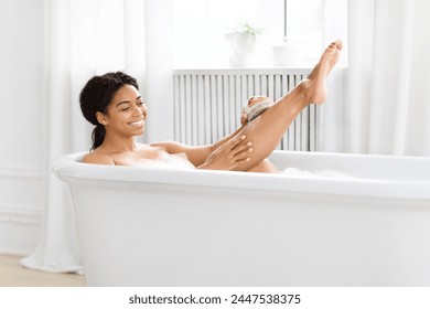 Playful young African American woman scrubbing her legs with brush while enjoying a bubble bath at home - Powered by Shutterstock