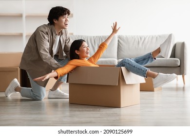 Playful Young Asian Couple Having Fun In Their Home On Relocation Day. Millennial Woman And Her Boyfriend Playing Together With Carton Box While Moving To New Apartment