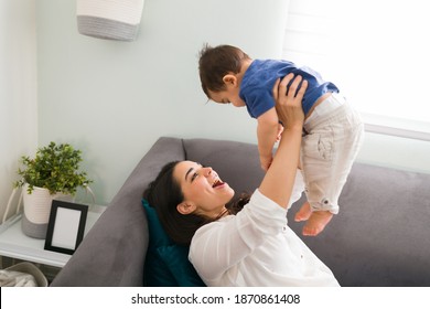 Playful Young Caucasian Mother Lying On Couch In Living Room Holding Up Her Baby Boy