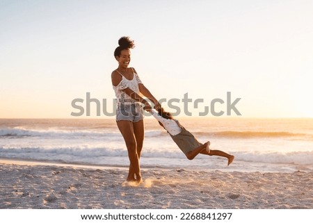 Playful young mother with laughing daughter fooling around at tropical beach during sunset. Young mother having fun while swinging her little black girl at sunset. Mother and daughter playing together