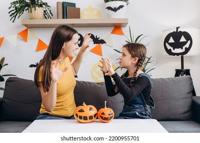 Playful Young Mother Sitting On Comfy Couch In Living Room With Cute Little Daughter Tells Spooky Halloween Stories. Funny Mom And Girl Kid Have Fun At Home. Celebration And Autumn Holiday Concept