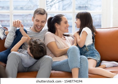 Playful Young Parents Have Fun Tickle Cute Small Kids Daughter And Son Enjoy Weekend At Home, Happy Mom And Dad Sit Relax On Couch Play With Smiling Little Kids, Spend Family Day Together